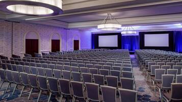 Un salón de conferencias con filas de sillas, una pantalla grande y una zona de escenario.