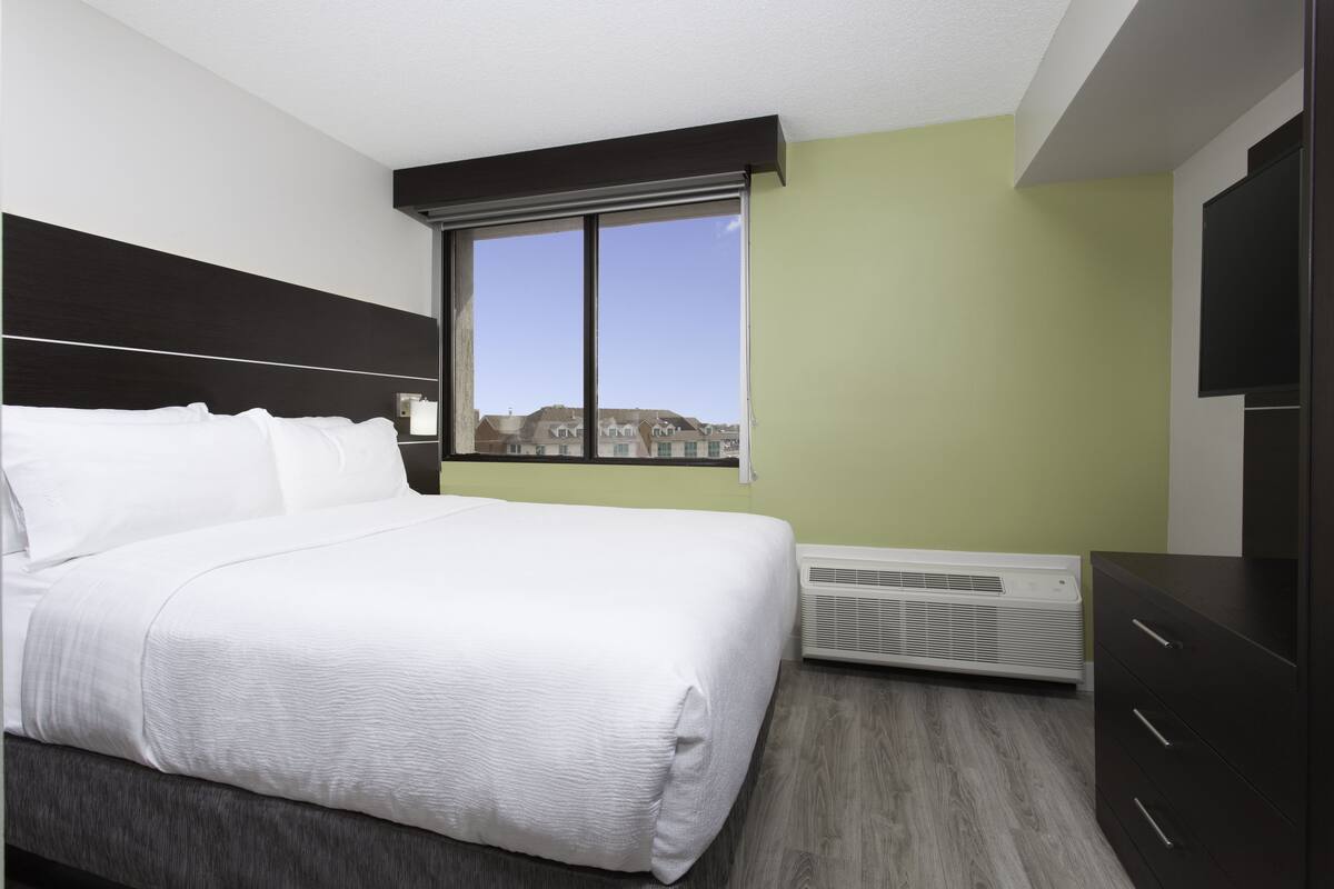 Ein modernes Hotelzimmer mit einem großen Bett, einem Fenster mit Blick auf Gebäude, einem Wand-Fernseher und einer eingebauten Klimaanlage.