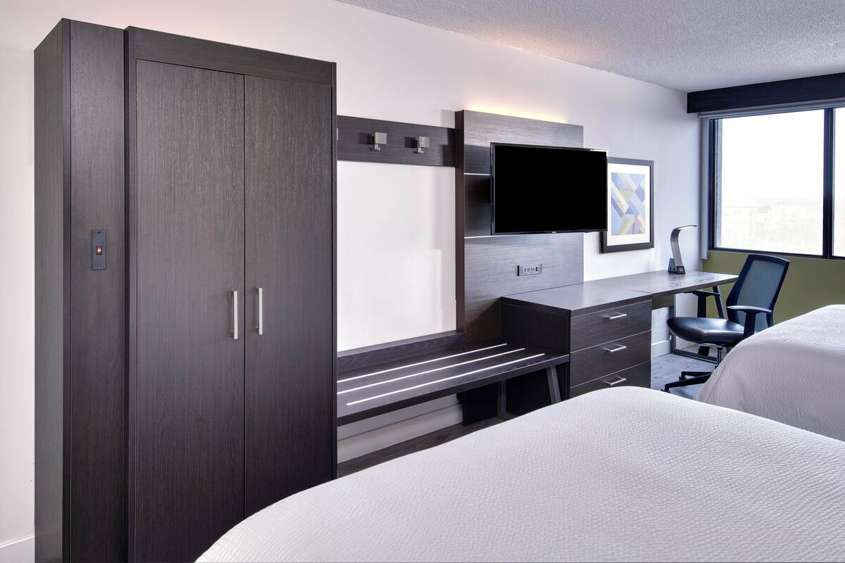 Ein modernes Hotelzimmer mit großem Kleiderschrank, einem Schreibtisch, einem Flachbildfernseher, einem Sessel und einem Bett.