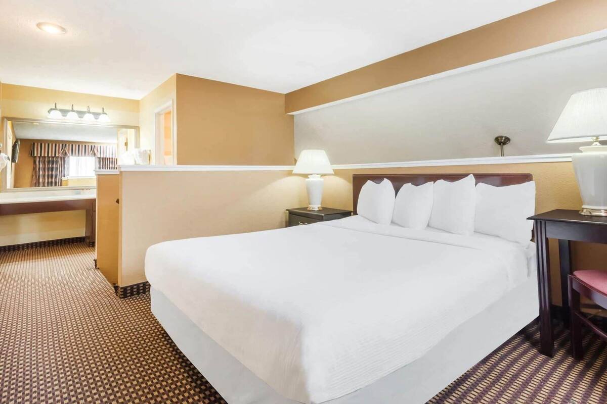 Une chambre d’hôtel avec un grand lit, une table de chevet avec une lampe, un miroir et une salle de bain visible au fond.