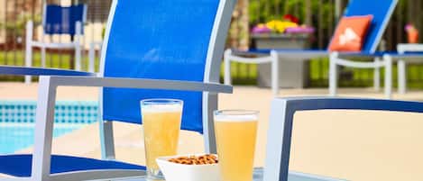 Un área junto a la piscina con sillas azules y una mesa. Sobre la mesa hay dos vasos de una bebida amarilla y un cuenco con aperitivos.