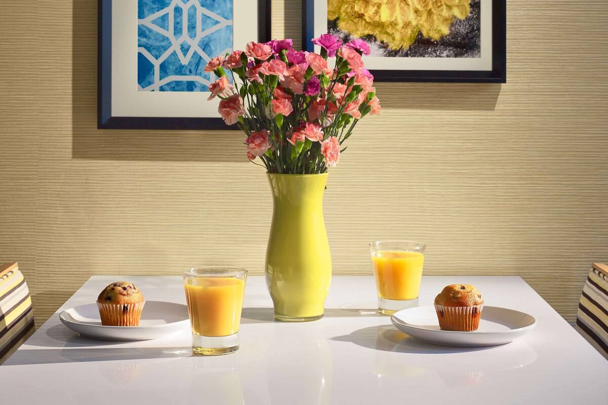 Una mesa con un jarrón amarillo de flores rosadas, dos muffins y dos vasos de jugo de naranja.
