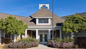 A building with a peaked roof, a flagpole with an American flag, and a small garden with pink flowers. - Sonesta ES Suites Cincinnati - Sharonville East (Sharonville)