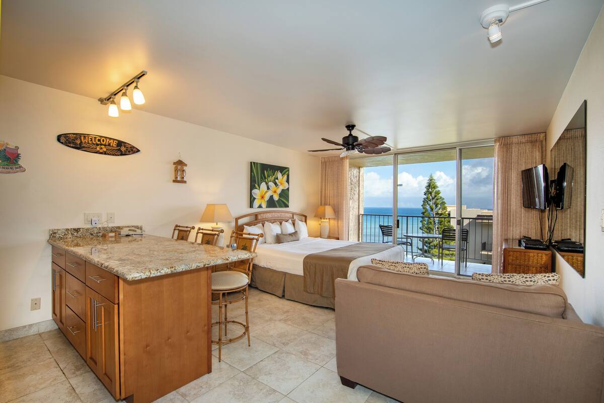 A hotel room with a kitchen area, a bed, a sofa, and a balcony with a view of the sea.