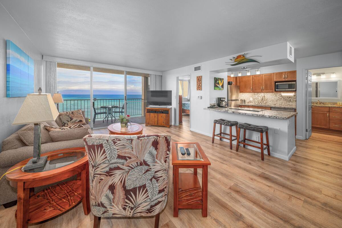 A spacious living area with a large window overlooking the ocean, a kitchen with wooden cabinets, and a dining area with a round table and bar stools.
