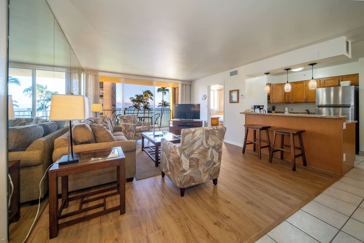 A spacious living area with a sofa, armchair, and coffee table, a kitchen with a refrigerator and bar stools, and a balcony with a view of palm trees.
