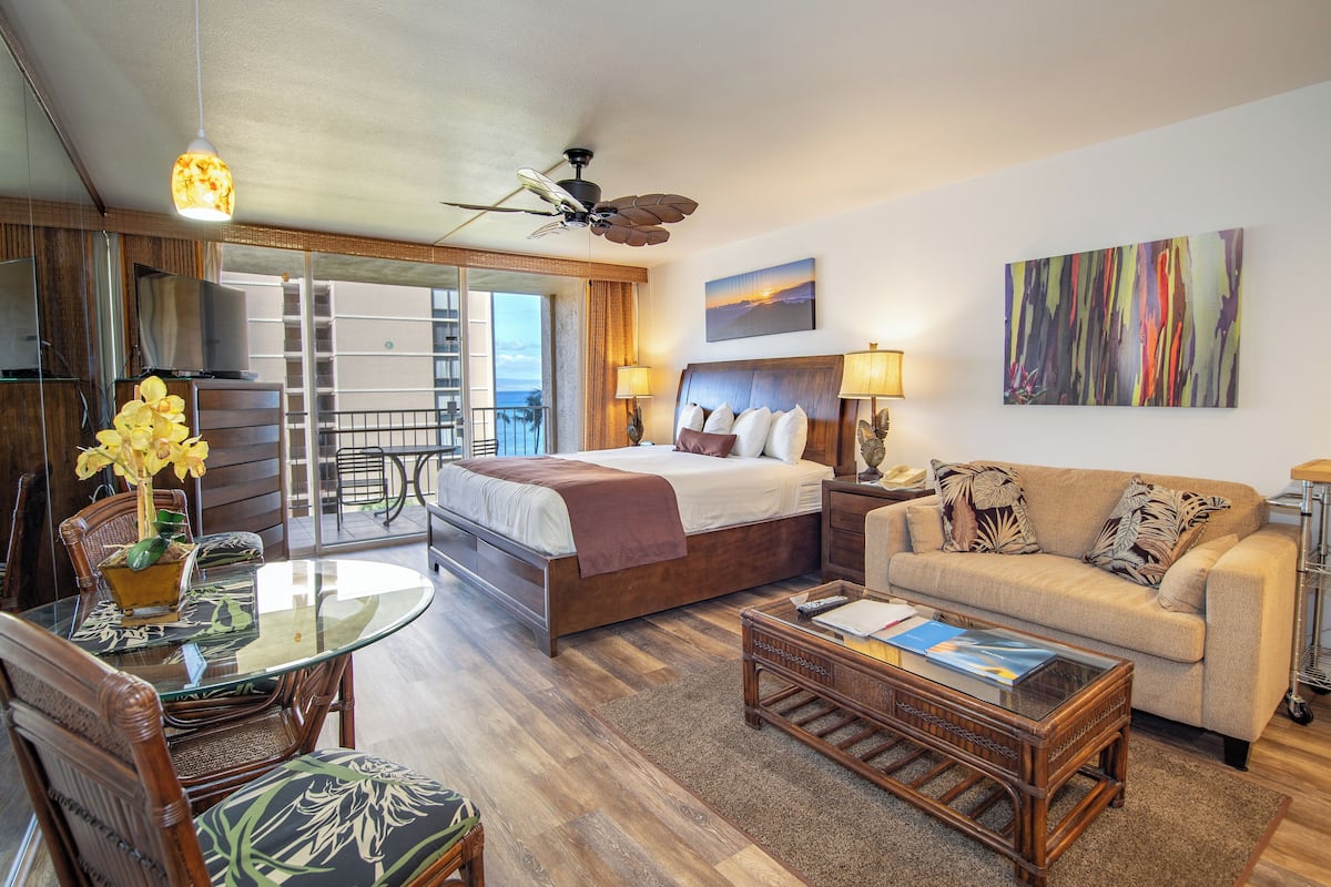 A hotel room with a bed, a sofa, a dining table with chairs, a balcony, and a ceiling fan.