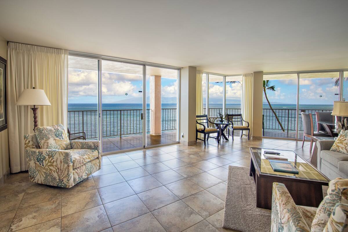A spacious living room with a large sliding glass door leading to a balcony with a view of the ocean. There are two armchairs and a coffee table in the room.