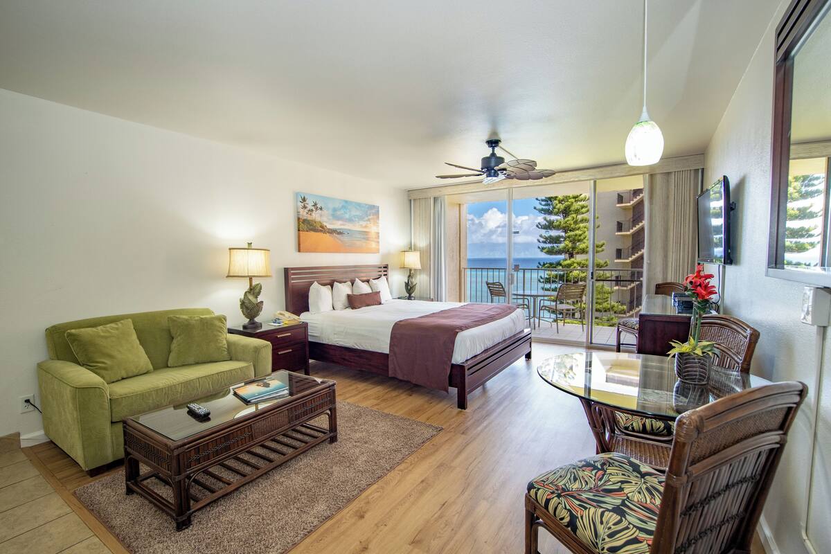 A hotel room with a bed, a green sofa, a dining table with chairs, a ceiling fan, and a view of the ocean through large windows.