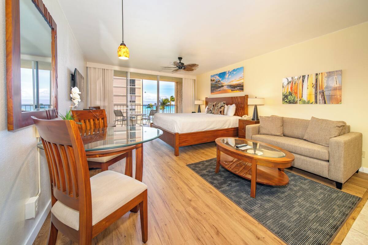 A hotel room with a bed, a sofa, a dining table with chairs, a glass coffee table, and a balcony with a view of the ocean.