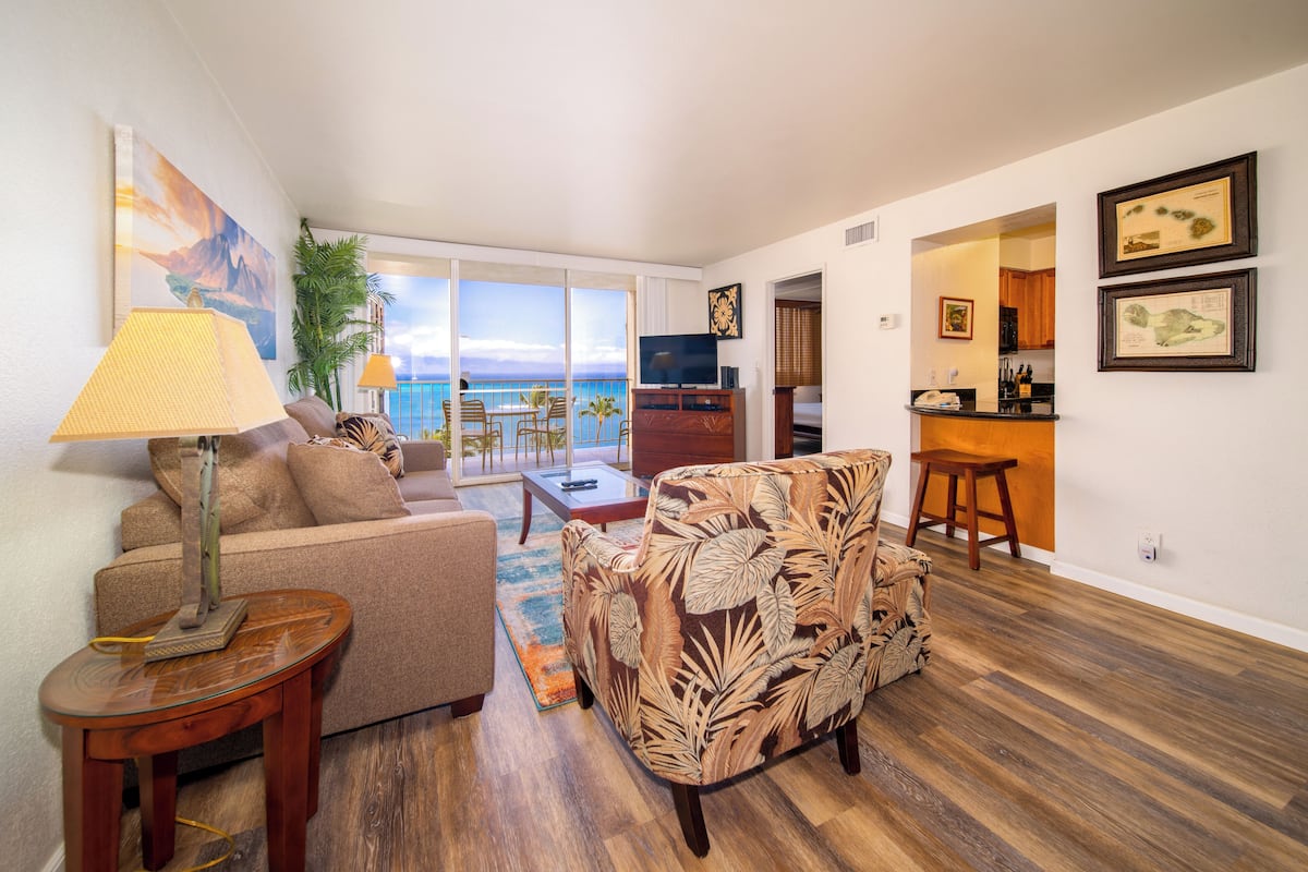 A living room with a sofa, armchair, and coffee table, a TV, and a view of the ocean.