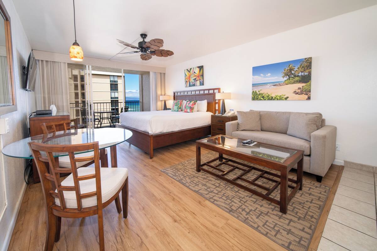 A hotel room with a bed, a sofa, a dining table with chairs, a coffee table, a ceiling fan, and a large window with a view of the beach.
