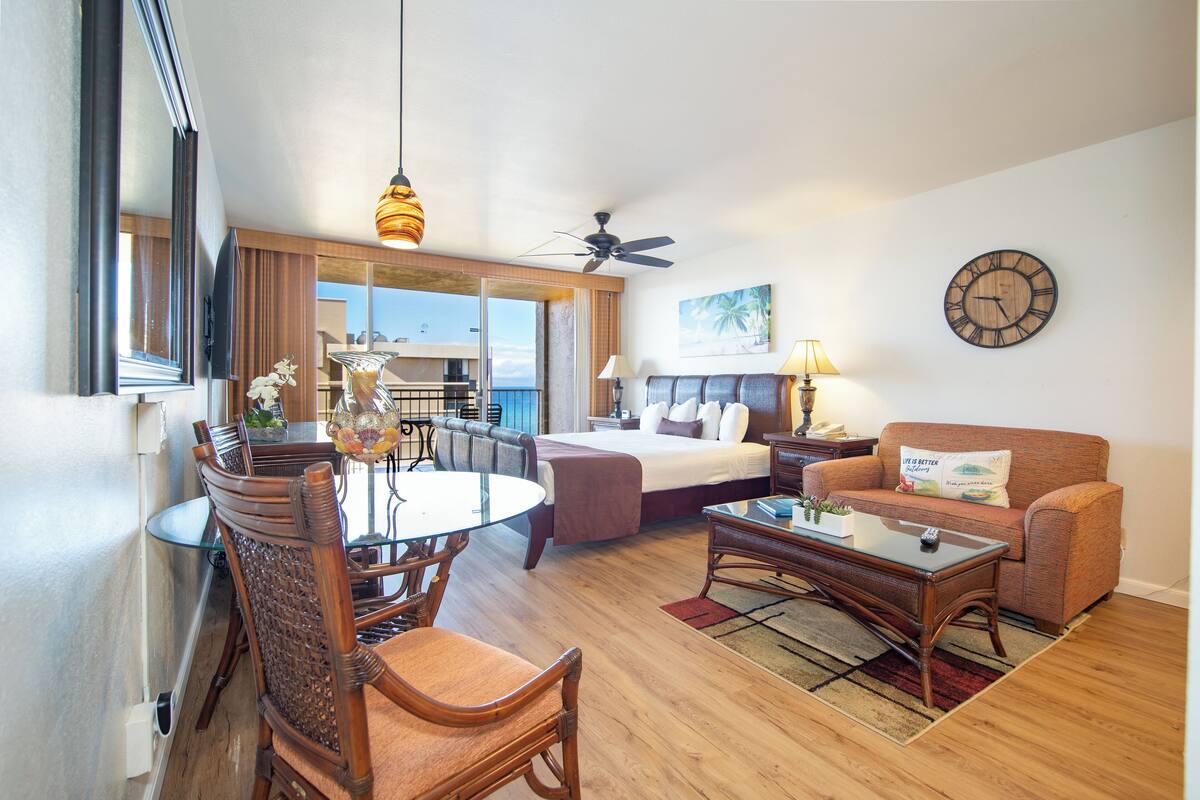 A hotel room with a bed, a sofa, a coffee table, a dining table with chairs, a ceiling fan, a clock, and a window with curtains.