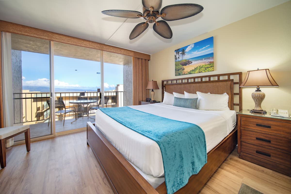 A hotel room with a large bed, a ceiling fan, a balcony with a view of the beach, and a painting on the wall.