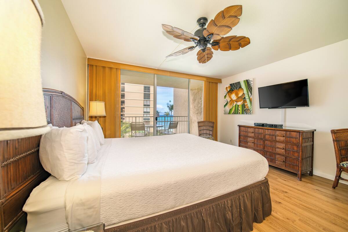 A hotel room with a large bed, a ceiling fan, a TV, a dresser, and a balcony with a view of the city.