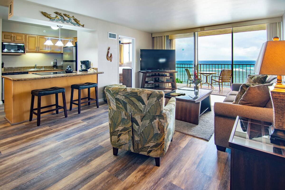 A modern living area with a kitchen, a large flat-screen TV, and a balcony with a view of the ocean.