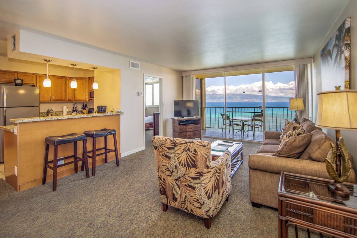 A hotel room with a kitchen, a living area with a sofa and armchair, a balcony with a view of the ocean, and a television.