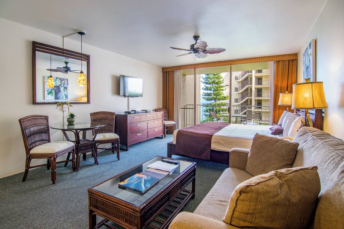 A hotel room with a bed, a sofa, a desk with a computer, a TV, a ceiling fan, and a view of the outside balcony.