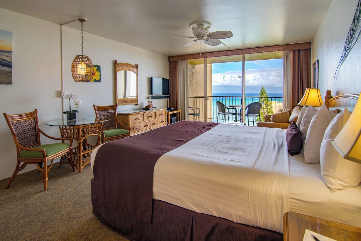 A hotel room with a large bed, a desk with a television, a balcony with a view of the ocean, and a dining area with chairs.