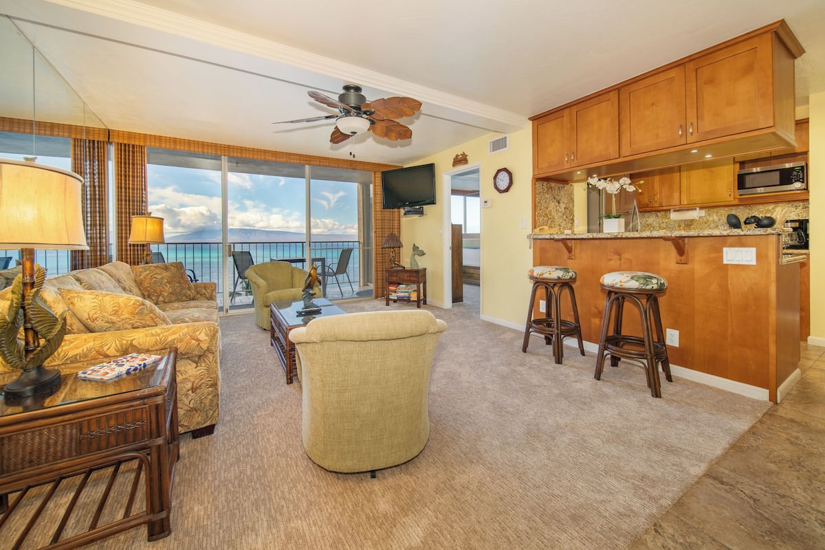 A spacious living room with a sofa, armchairs, and a coffee table. There is a balcony with a view of the ocean. The kitchen has wooden cabinets and a microwave.