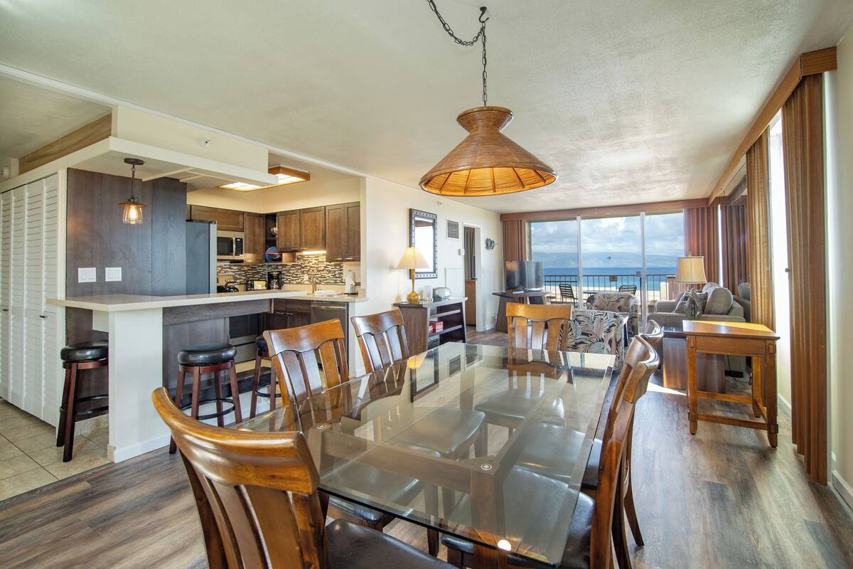 A spacious dining area with a glass table and wooden chairs, a kitchen with dark wood cabinets, and a view of the sea through large windows.