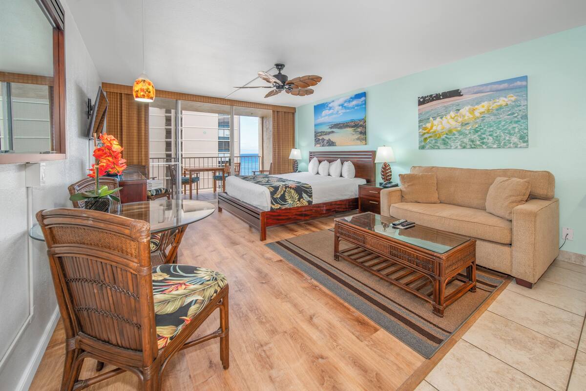 A hotel room with a bed, a sofa, a dining table, and a balcony with a view.