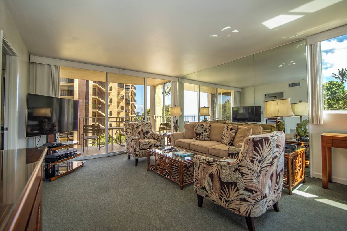 A spacious living room with a large flat-screen TV, comfortable seating, a glass coffee table, and a view of a building with balconies.