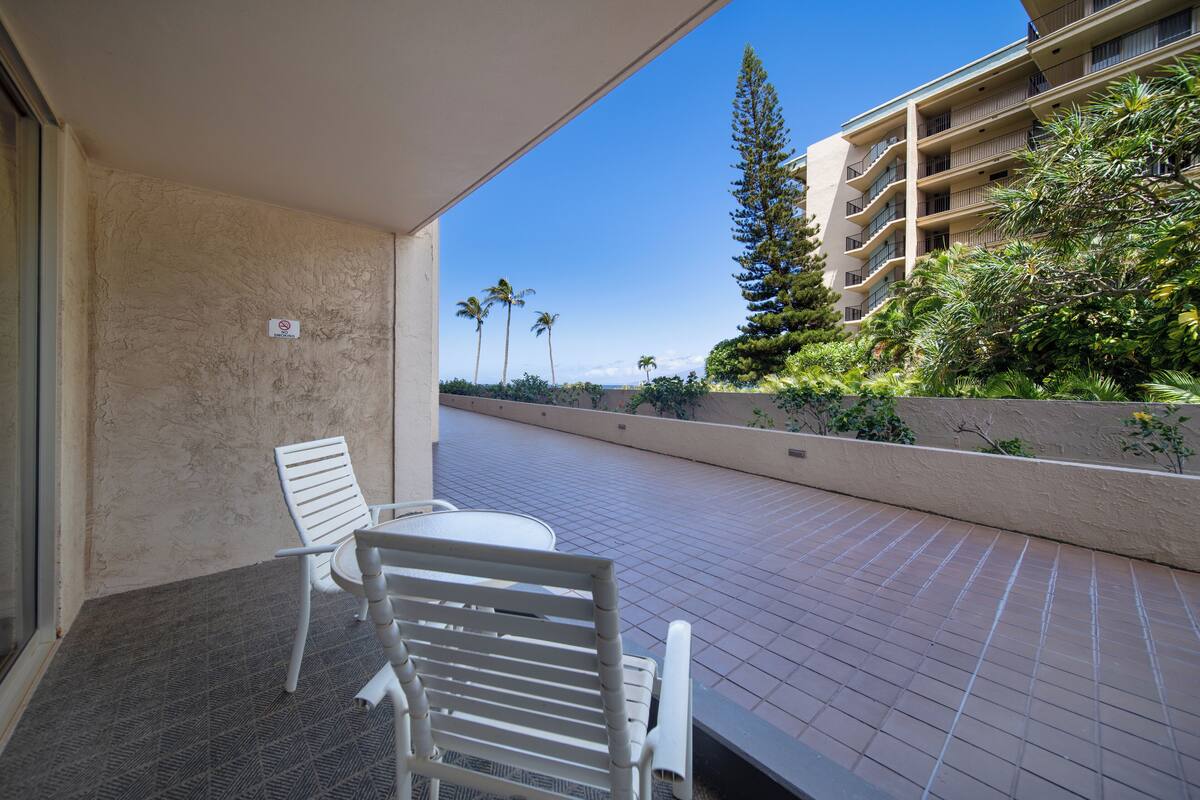 A balcony with two white chairs and a table, overlooking a paved walkway and a building with balconies and greenery.