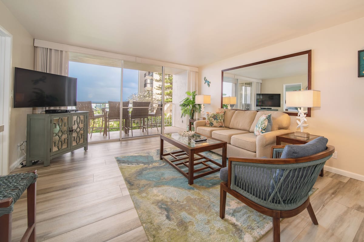 A spacious living room with a large flat-screen TV, a sofa, a coffee table, and a chair. There is a balcony with a view of the city.