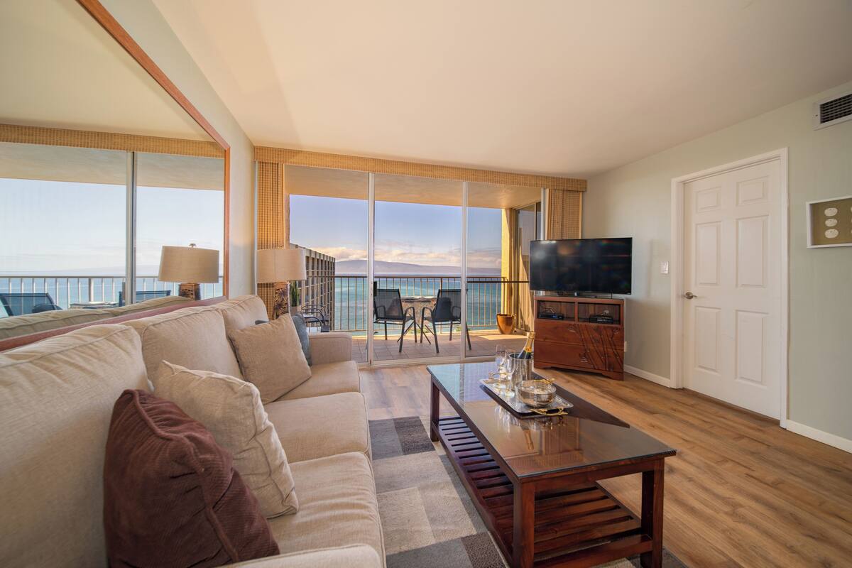 A spacious living room with a large sofa, a coffee table, and a TV. There is a balcony with a view of the ocean.