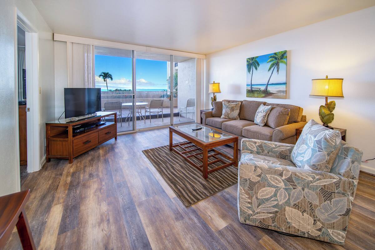 A living room with a TV, sofa, and coffee table, a balcony with a view of the beach, and a palm tree painting on the wall.