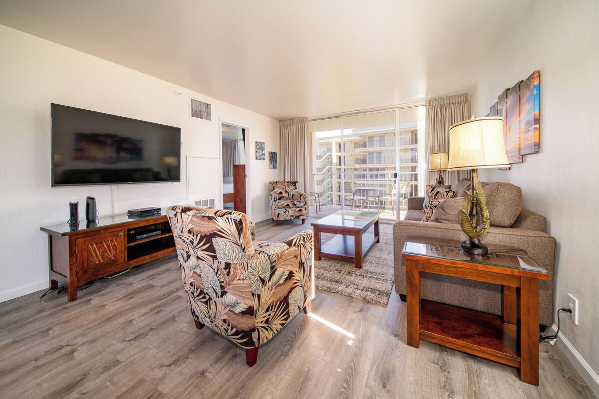 A modern living room with a flat-screen TV, a patterned armchair, a sofa, a glass-top coffee table, and a lamp.