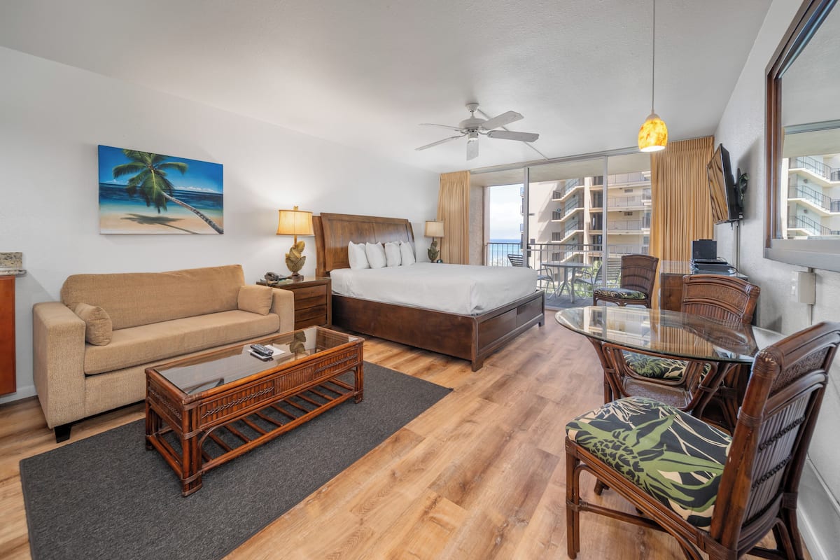 A hotel room with a bed, a sofa, a dining table with chairs, a ceiling fan, and a large window with a view of the city.