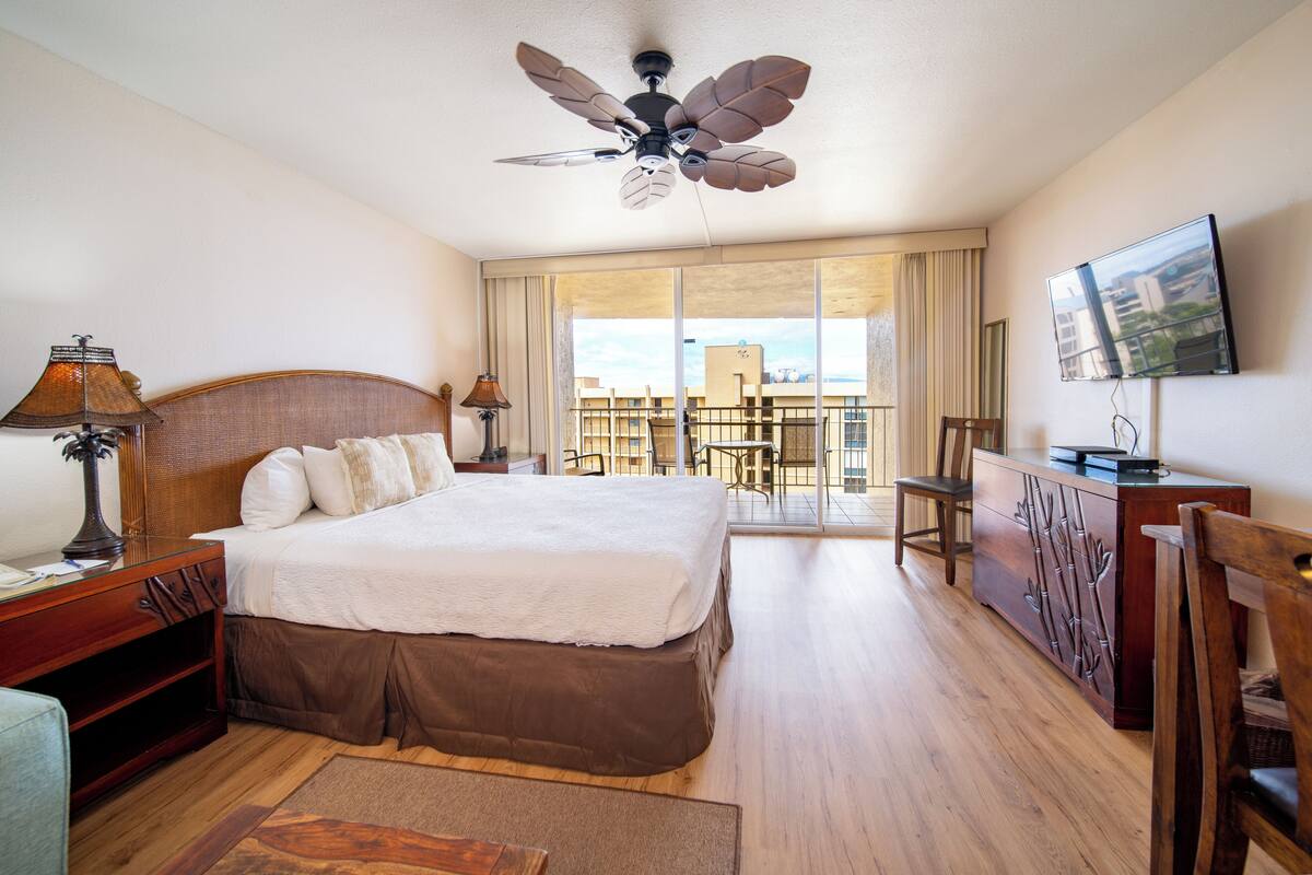 A hotel room with a large bed, wooden headboard, a ceiling fan, a TV, a desk with a lamp, and a view of the outside balcony.
