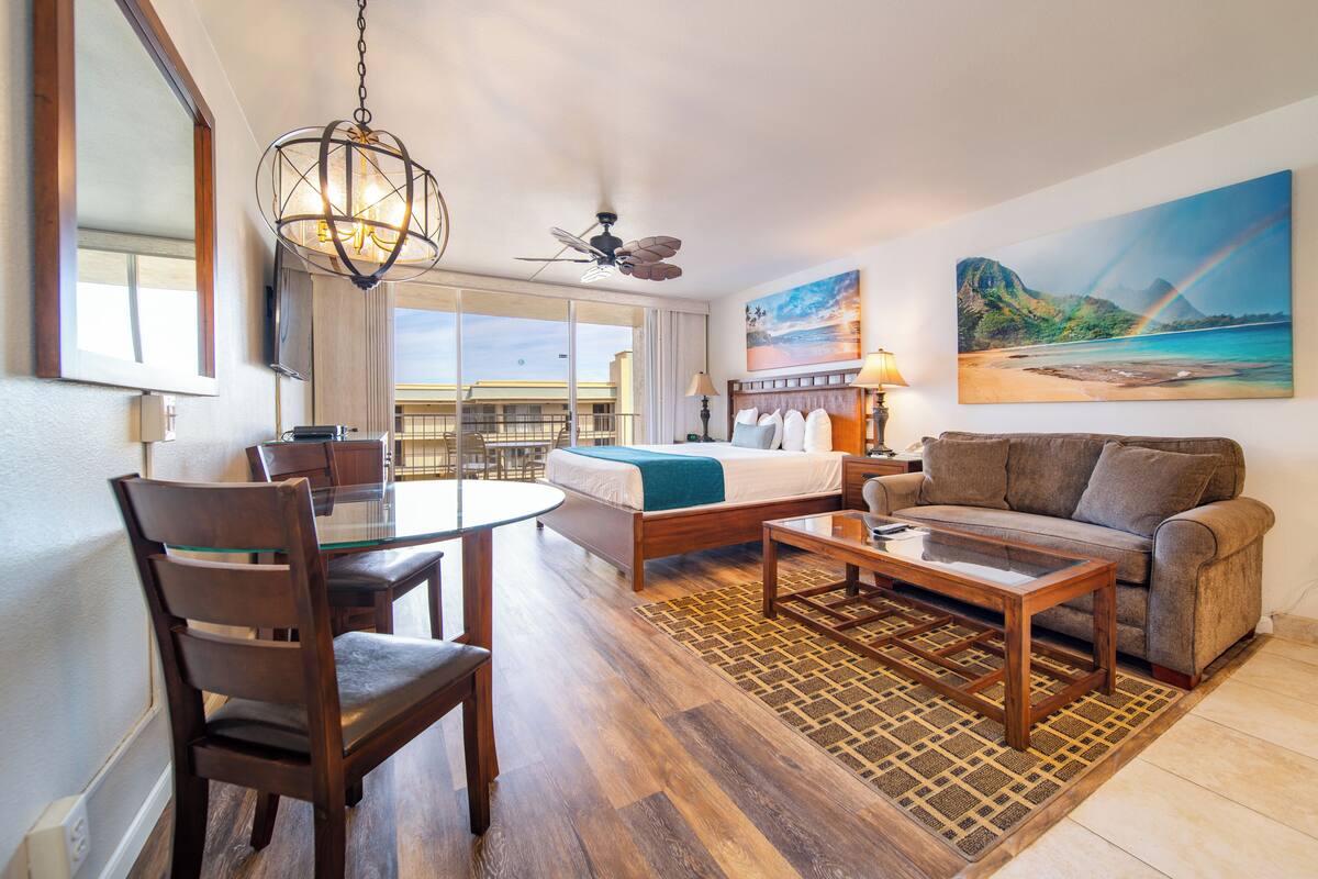 A hotel room with a bed, a sofa, a dining table with chairs, a chandelier, a ceiling fan, and a large window with a view of the beach.