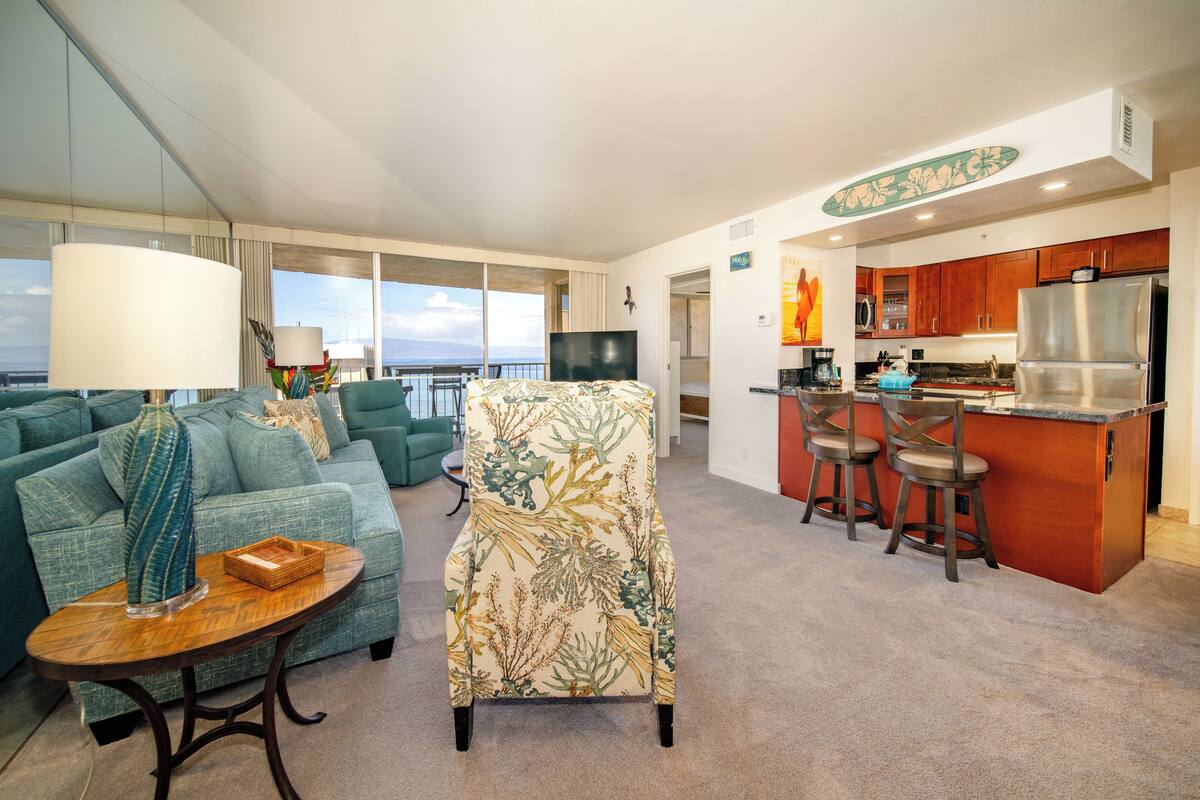 A modern living room with a sofa, a patterned armchair, a small table, and a kitchen area with a bar and stools.