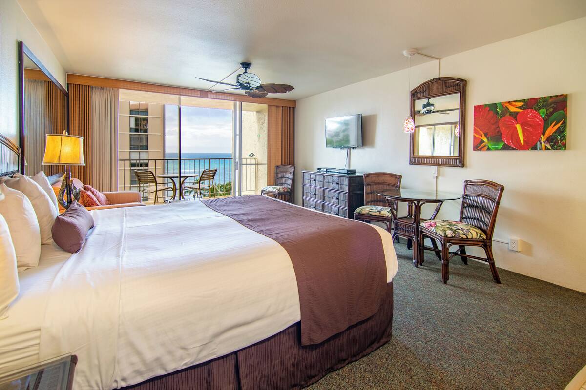 A hotel room with a large bed, a desk with a chair, a television, and a balcony with a view of the ocean.