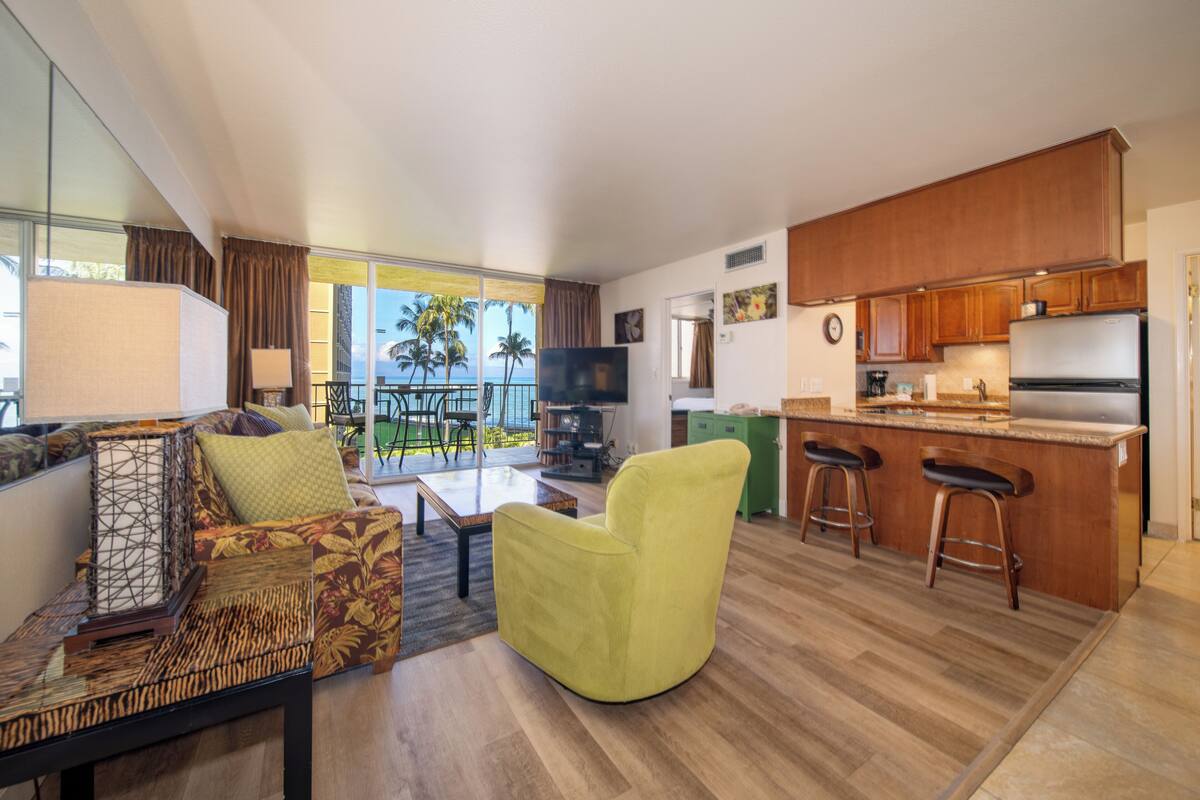 A modern living room with a sofa, a yellow armchair, and a coffee table. There is a kitchen area with a refrigerator, a sink, and a bar counter with stools. The room has large windows with a view of palm trees.