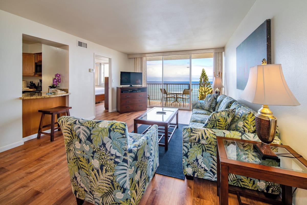 A living room with a sofa, coffee table, and TV, a balcony with a view, and a kitchen area.
