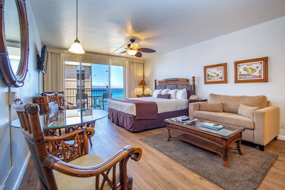 A hotel room with a bed, a sofa, a dining table with chairs, a ceiling fan, and a balcony with a view of the ocean.