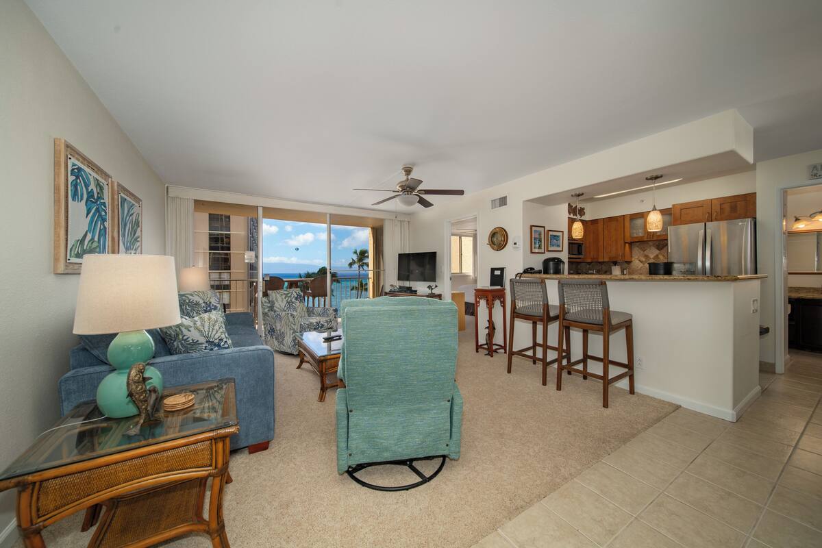 A spacious living area with a sofa, chairs, and a dining table. A kitchen is visible with wooden cabinets and a refrigerator.