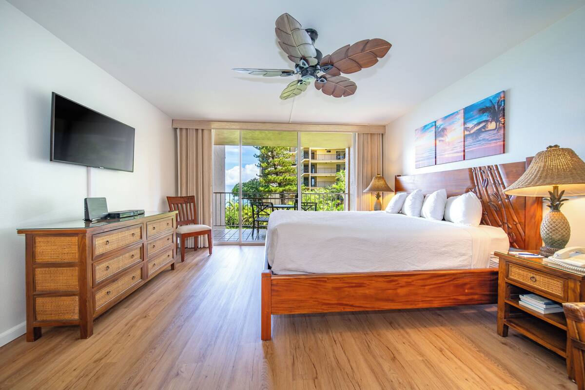 A hotel room with a wooden bed, a desk with drawers, a flat-screen TV, a ceiling fan, a window with a view of greenery, and a painting on the wall.