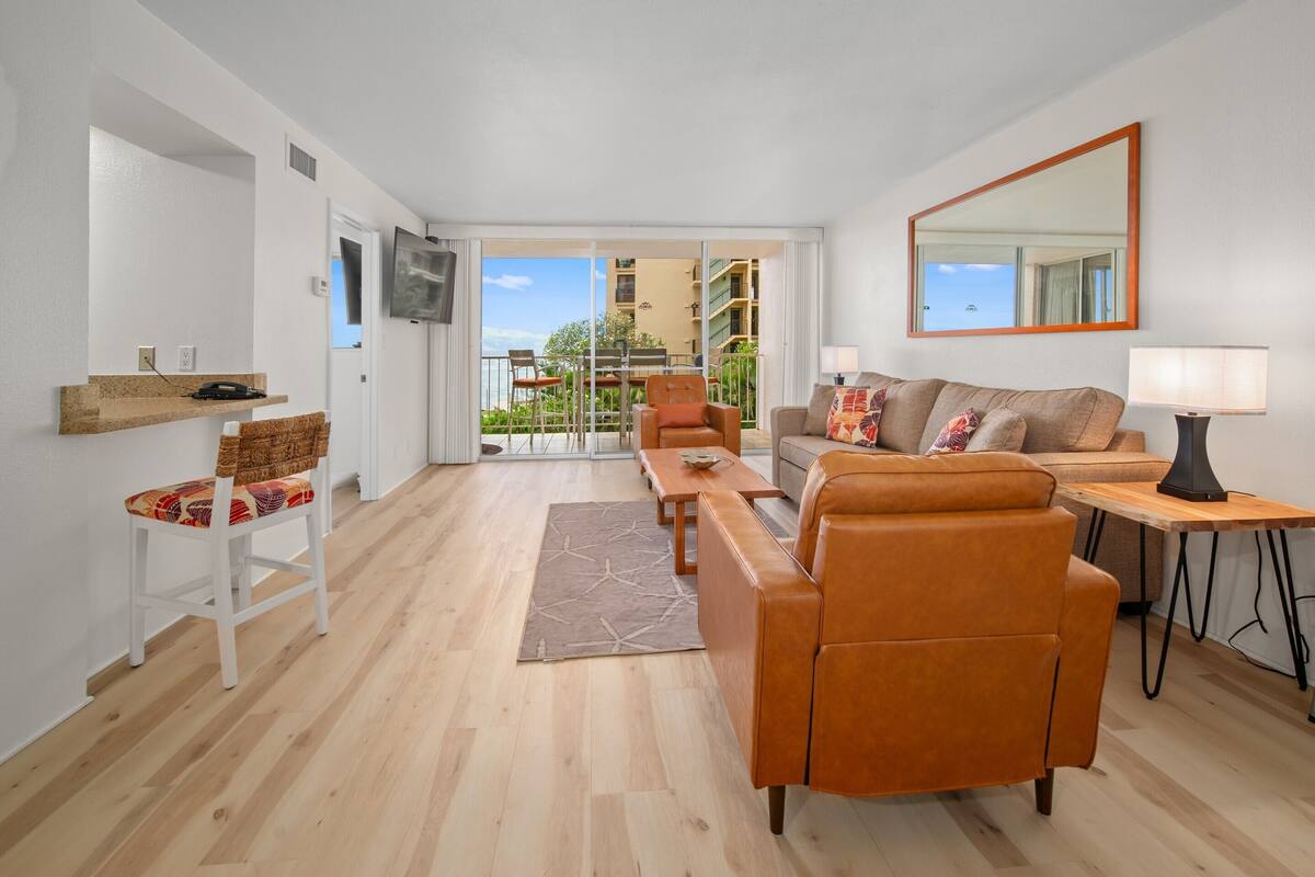A modern living room with a sofa, armchair, and a dining table. There is a TV mounted on the wall, and a balcony with a view of buildings.