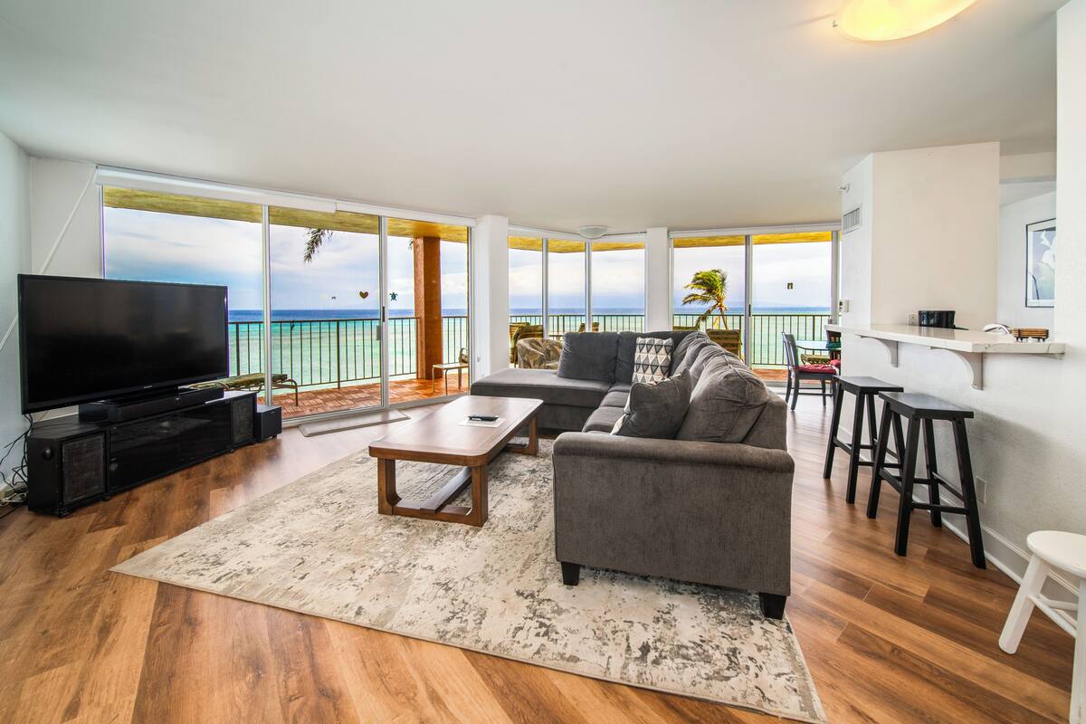 A spacious living room with a large flat-screen TV, comfortable seating, and a view of the ocean through large windows.