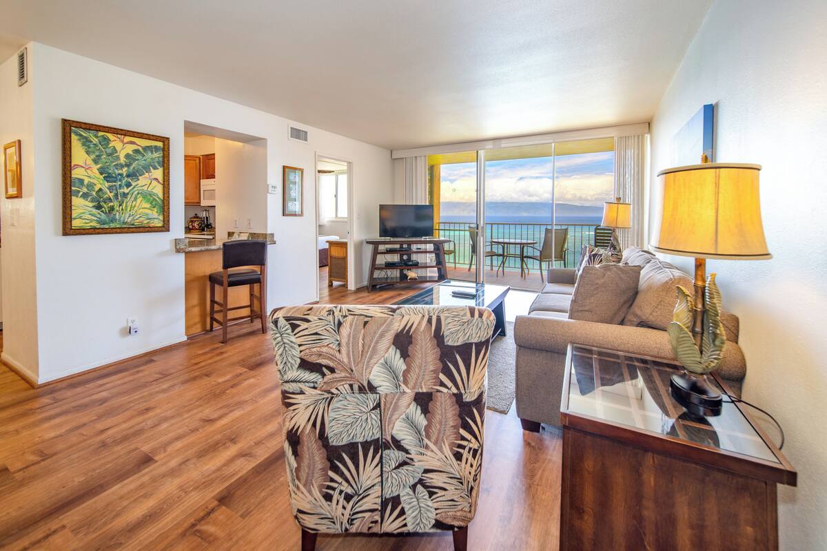A spacious living room with a large window offering a view of the ocean, a television, a sofa, and a patterned armchair.