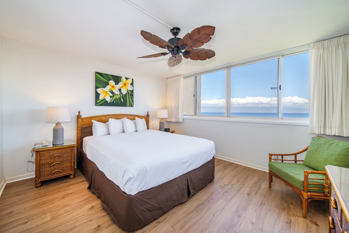 A hotel room with a large bed, a wooden nightstand, a green chair, a ceiling fan, and a window with a view of the ocean.