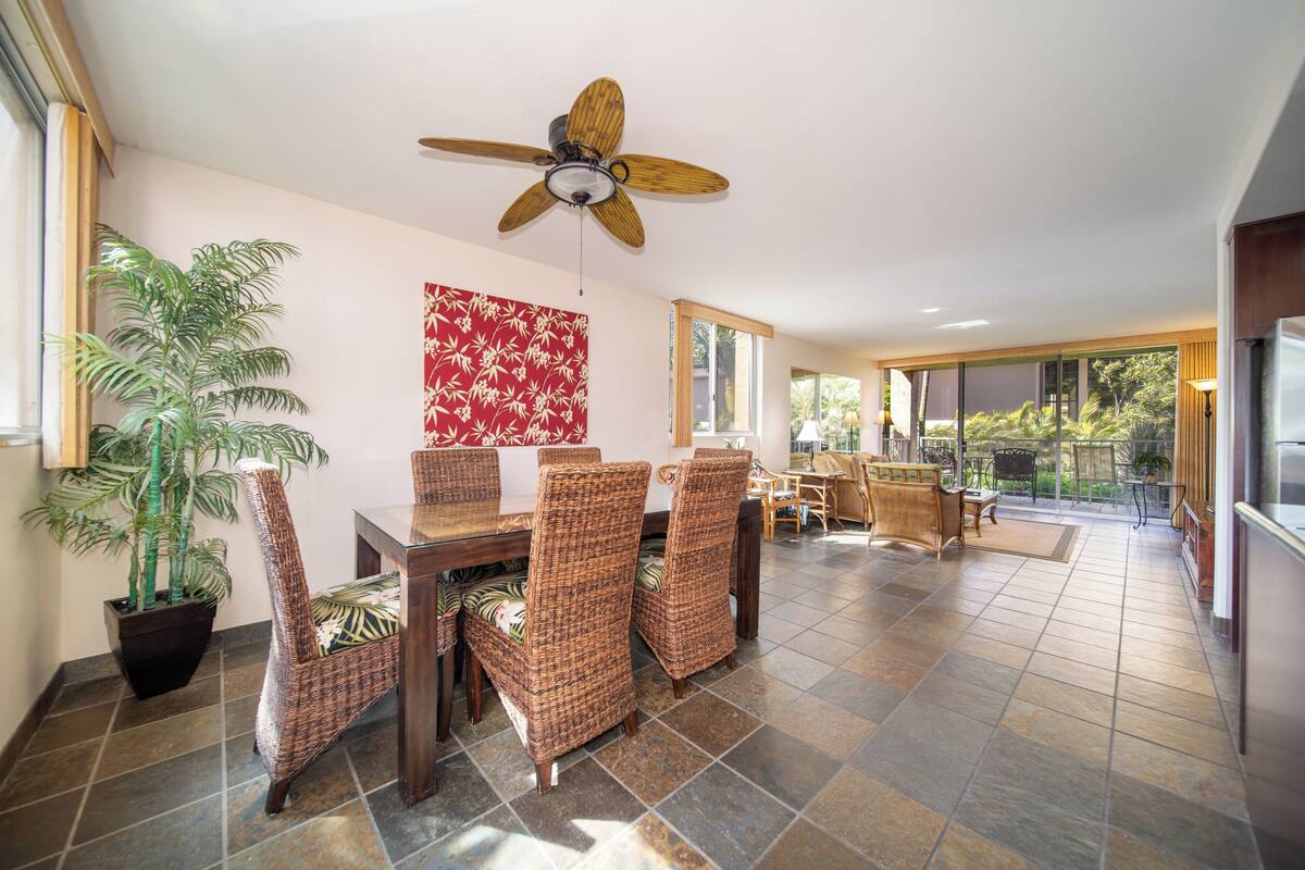 A dining area with wicker chairs, a wooden table, a ceiling fan, and a large window offering a view of the outdoors.
