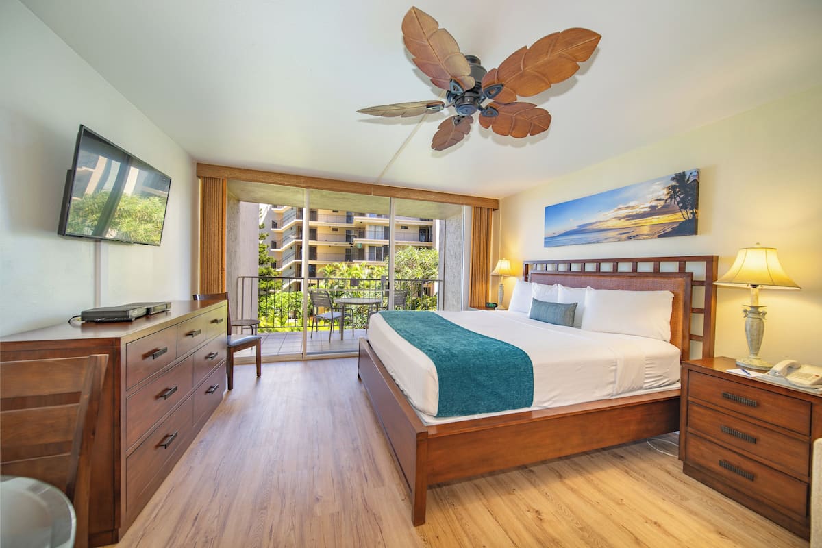 A hotel room with a wooden bed, a ceiling fan, a balcony with a view of a building, and a painting on the wall.
