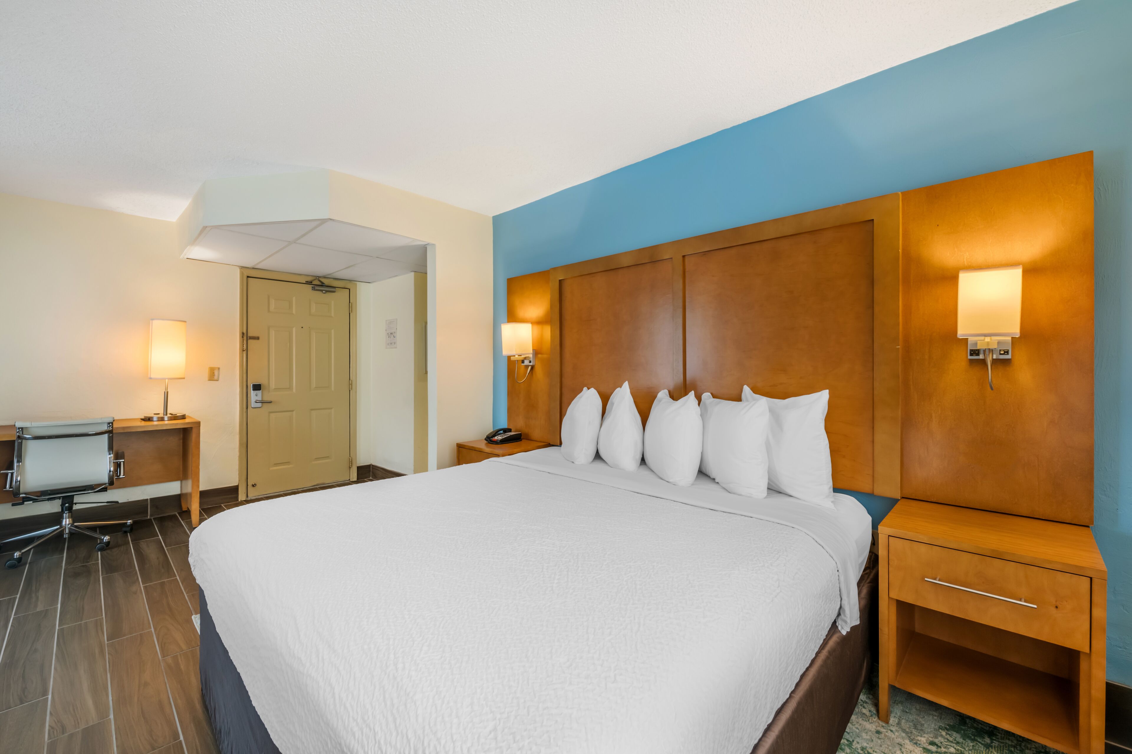 A hotel room with a large bed, two bedside tables with lamps, a desk with a chair, and a door leading to another room.