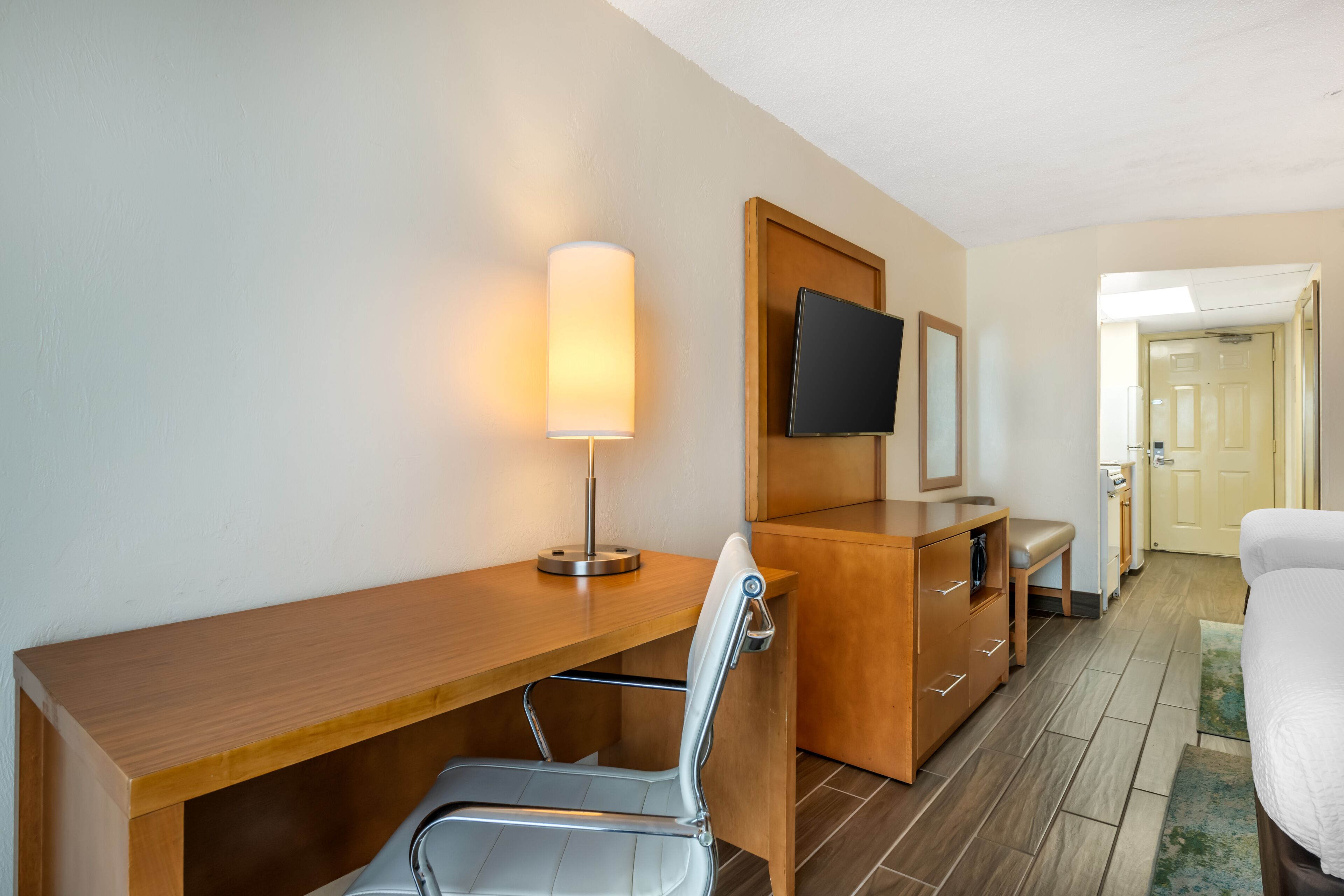 A hotel room with a wooden desk, a chair, a television mounted on the wall, and a bed with a white comforter.